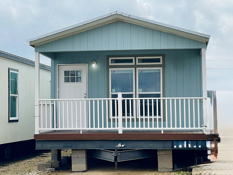 Exterior of Landmark RV Park Model home, Light blue on elevated blocks with a front deck and white railings.
