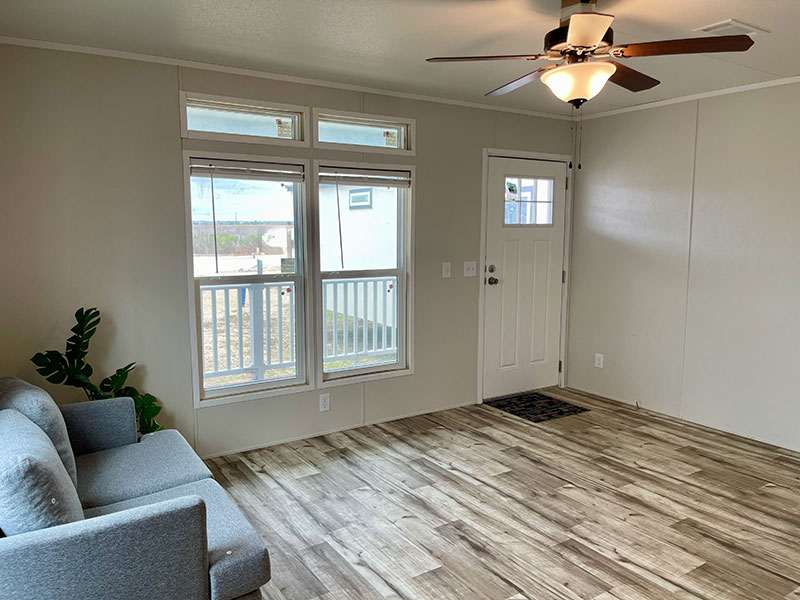 Landmark RV Park Model home, A living room with a gray sofa, wooden floor, large windows, a ceiling fan, and a white door.