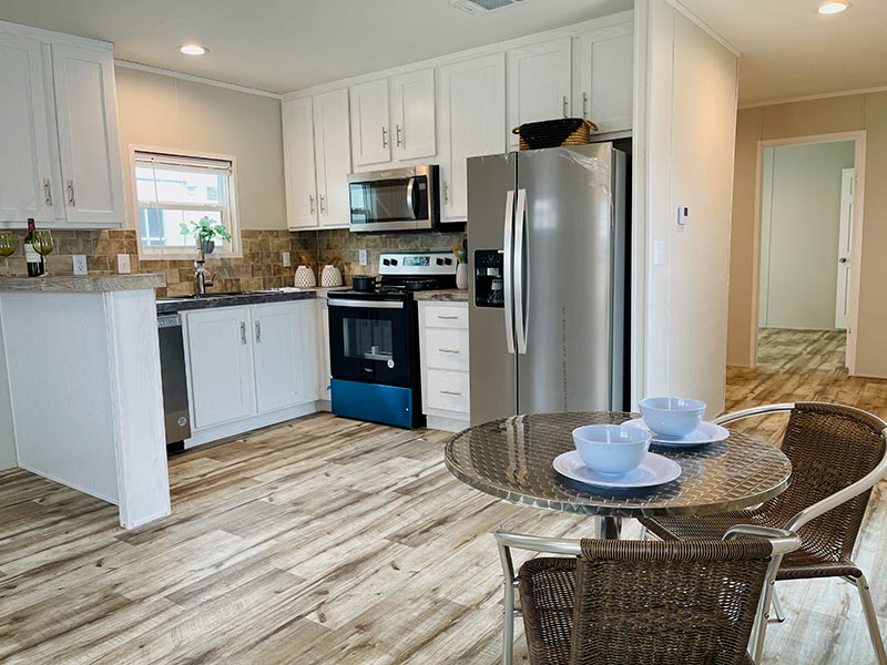 Landmark RV Park Model home, Modern kitchen with white cabinets, stainless steel appliances and a glass dining table with wicker chairs.