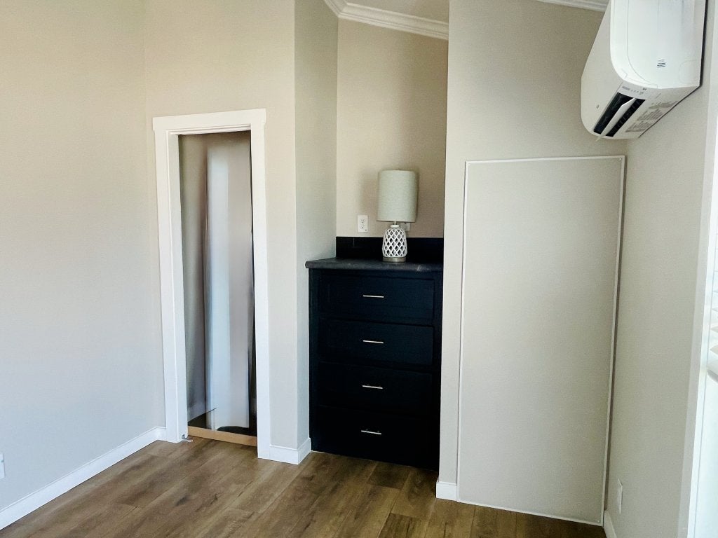 Hill Country RV Park Model tiny home modern Bedroom featuring a black dresser with a lamp, a partially open closet door, and a mounted air conditioning unit, all on wooden flooring.