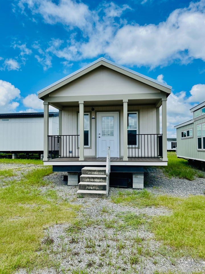 Crane RV Park Model tiny home small, light-colored with a gable roof and a covered porch, set against a blue sky with clouds. Concrete steps lead up to the porch, surrounded by gravel and patches of grass.