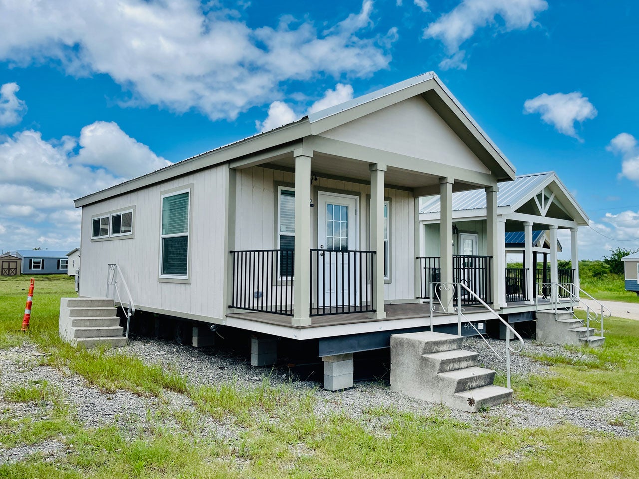 Crane RV Park Model tiny home modern factory-built with a gabled roof and a front porch, surrounded by grassy and gravel terrain under a blue sky.