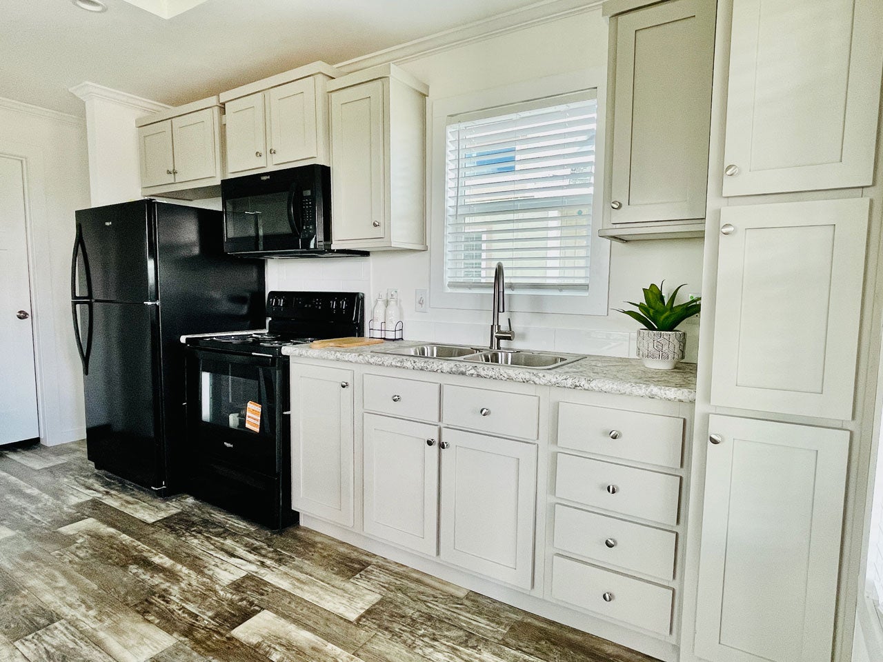 Crane RV Park Model tiny home modern kitchen featuring black appliances, white cabinetry, a dual basin sink, and a decorative green plant on the countertop, with wooden flooring.