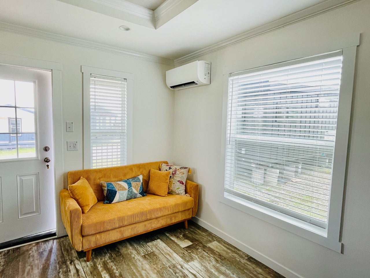Crane RV Park Model tiny home cozy corner of a room featuring a mustard corduroy sofa with patterned cushions, a white door with a glass panel, two windows with blinds, and an air conditioner mounted on the wall.
