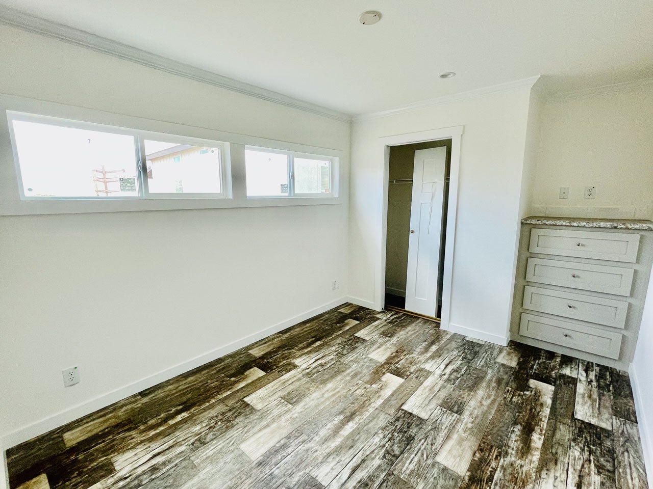 Crane RV Park Model tiny home Bright, minimalistic room featuring narrow windows, a gray dresser, and a closet door, with textured wooden flooring.