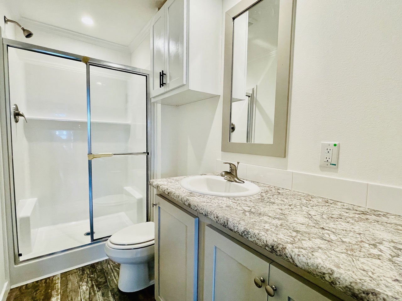 Crane RV Park Model tiny home modern bathroom with a glass-enclosed shower, white toilet, and granite countertop sink. A rectangular mirror is mounted above the sink.