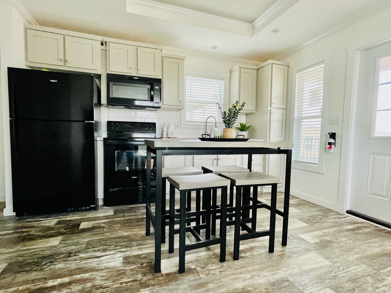 Crane RV Park Model tiny home modern kitchen with a black refrigerator, oven, and microwave, featuring a stone kitchen island with three gray bar stools and two bright windows.