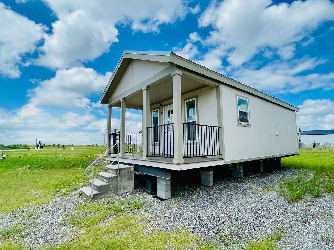 A modern Crane RV Park Model tiny home on blocks with a gabled roof and a porch, surrounded by grassy and gravel areas under a blue sky with clouds.