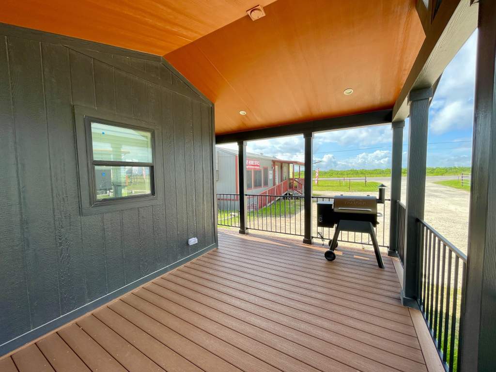 Exterior of Hill Country RV Park Model tiny home covered porch with wooden flooring, black walls, and an orange ceiling. A black grill is on the right, with a view of a red building and green field in the background.