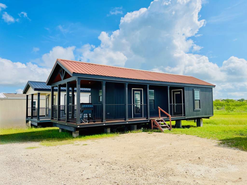 Exterior of Hill Country RV Park Model tiny home with dark siding and a red metal roof, featuring a porch with stairs, set against a blue sky with clouds.