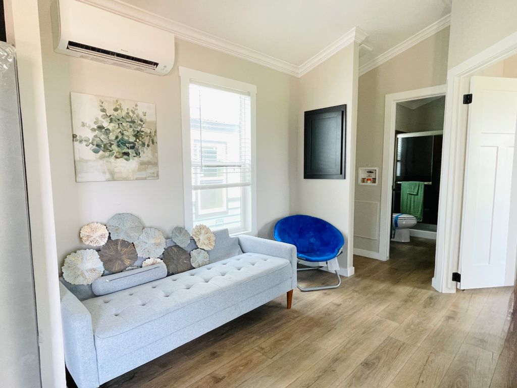 Hill Country RV Park Model tiny home Living Room featuring a light sofa with decorative pillows, a blue chair, and framed artwork, with visible air conditioning and access to a bathroom.