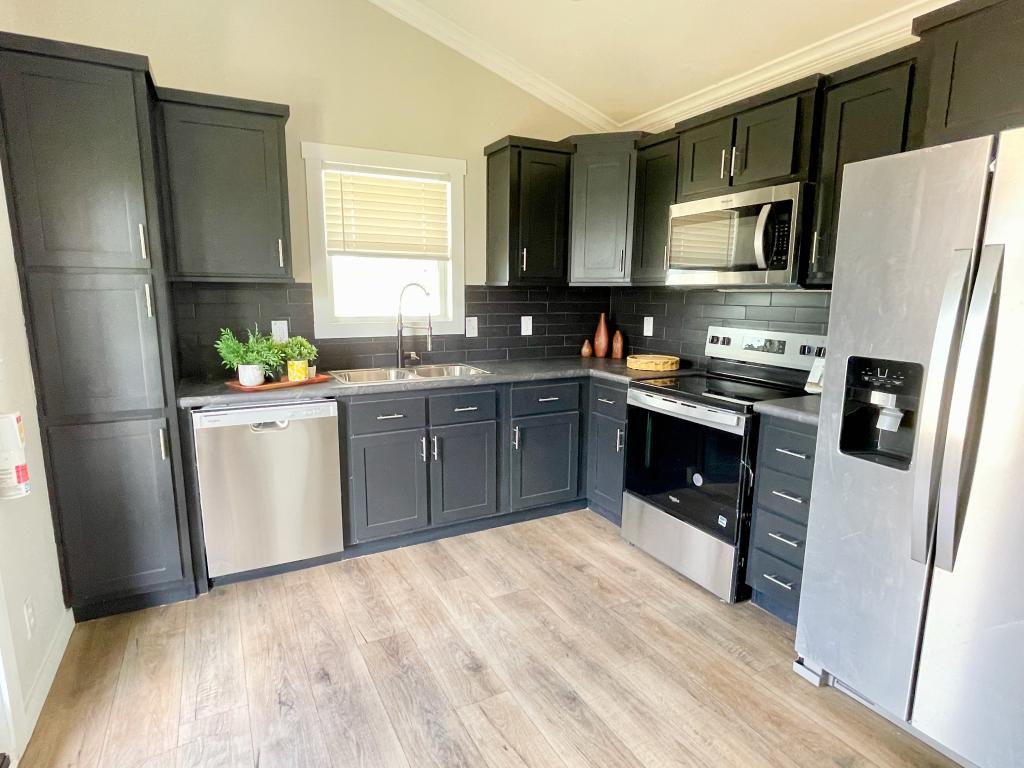 Hill Country RV Park Model tiny home Modern kitchen with black cabinets, stainless steel appliances, and a window above the sink, featuring light wood flooring.