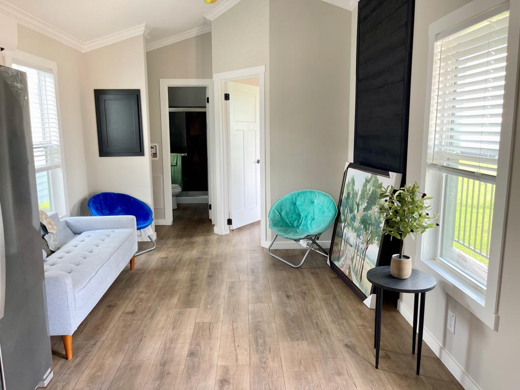 Hill Country RV Park Model tiny home a bright living space featuring a gray couch, blue and green accent chairs, a black table with a plant, and a large framed picture against the wall.