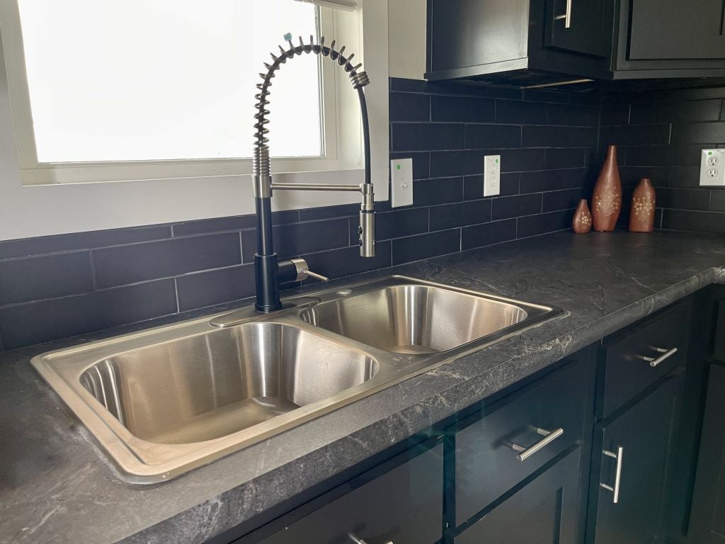 Hill Country RV Park Model tiny home modern kitchen featuring a stainless steel double sink, high-arc faucet, black granite countertop, and decorative vases against a black tiled backsplash.